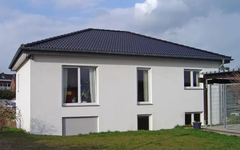 Moderner Bungalow mit weißer Putzfassade, dunkel gedecktem Satteldach und bodentiefen Fenstern, gelegen auf einem leicht abschüssigen Grundstück.