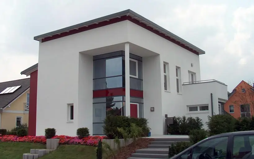 Modernes Einfamilienhaus mit flachem Satteldach, auffälliger Glasfassade im Eingangsbereich und rotem Akzentstreifen.