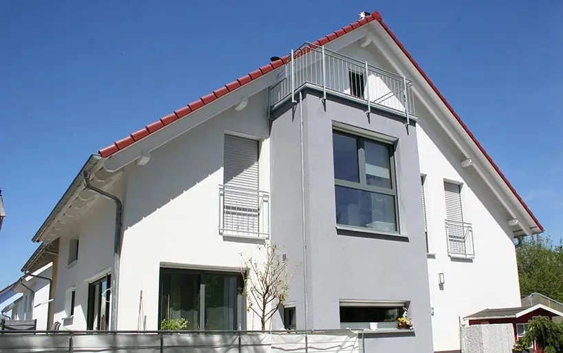Zweigeschossiges Einfamilienhaus mit weißer Fassade, rotem Satteldach und grauer Erkerfront mit großen Fenstern und kleinem Balkon im Dachgeschoss.