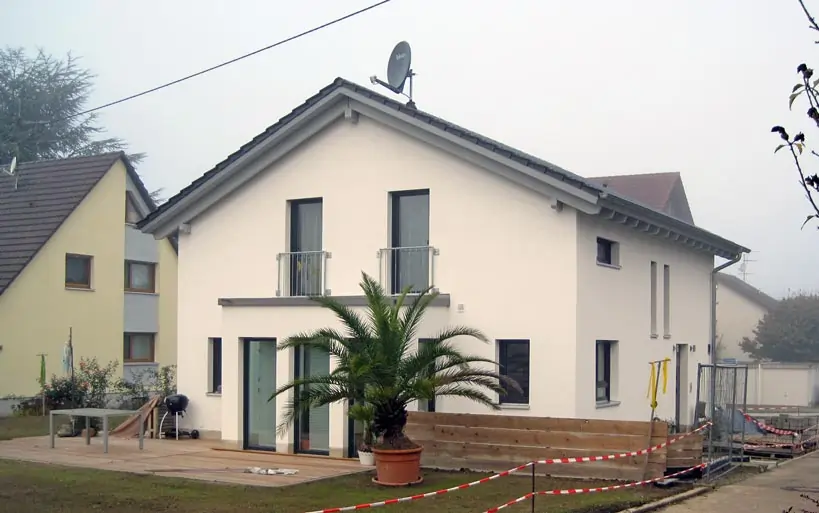 Modernes Einfamilienhaus mit weißer Putzfassade, grauem Satteldach und Balkonfenstern, davor eine große Palme in einem Blumentopf auf der Terrasse.