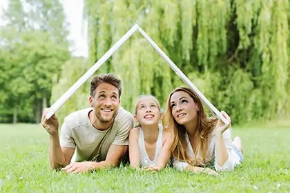 Glückliche Familie liegt auf einer Wiese unter einer symbolischen Dachform – Sinnbild für den Traum vom Eigenheim mit einem Hausbausatz.