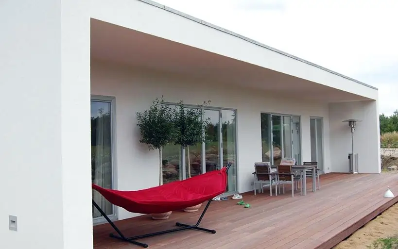 Moderner Flachdachbungalow mit großer überdachter Holzterrasse, roter Hängematte, Sitzgruppe und Glasfronten, die den Innenraum mit dem Garten verbinden.