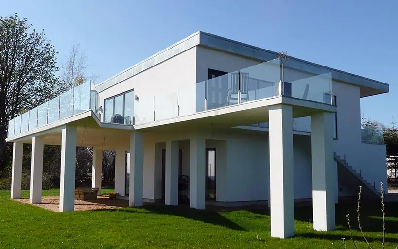 Zweigeschossiges Flachdachhaus mit großzügigem, umlaufendem Balkon und Glasgeländer auf Stützen, moderner Architekturstil.