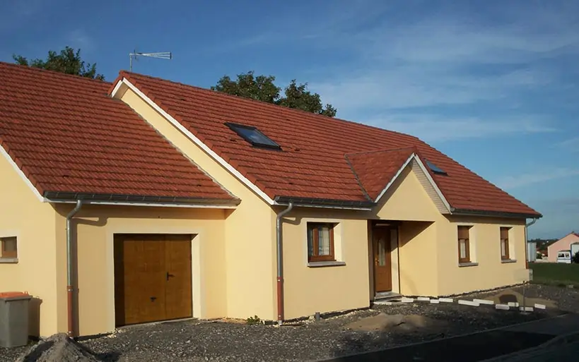 Einstöckiges Einfamilienhaus mit rotem Satteldach, integrierter Garage und gelber Fassade, aufgenommen während der Bauphase auf einem noch unbefestigten Grundstück.