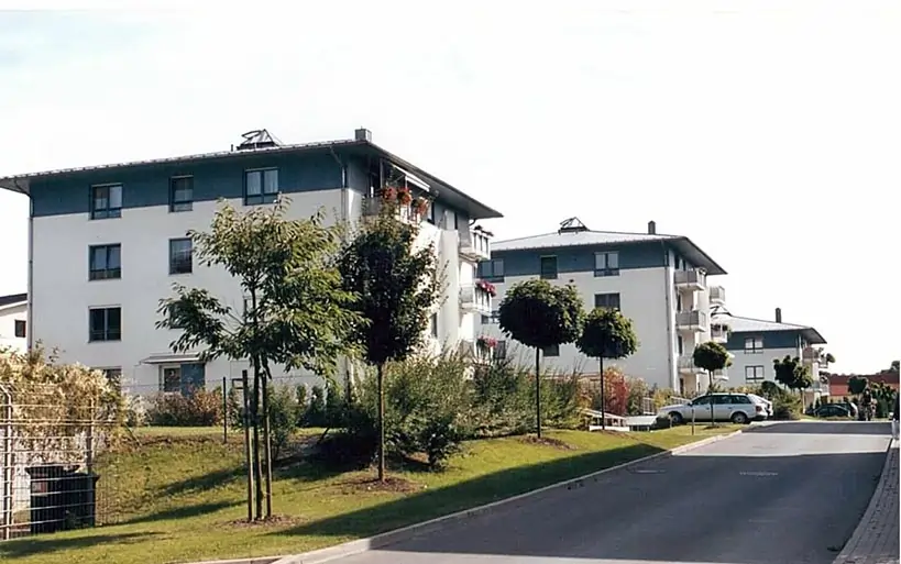 Reihe moderner Mehrfamilienhäuser mit Flachdach an einer begrünten Straße mit jungen Bäumen und geparkten Autos.