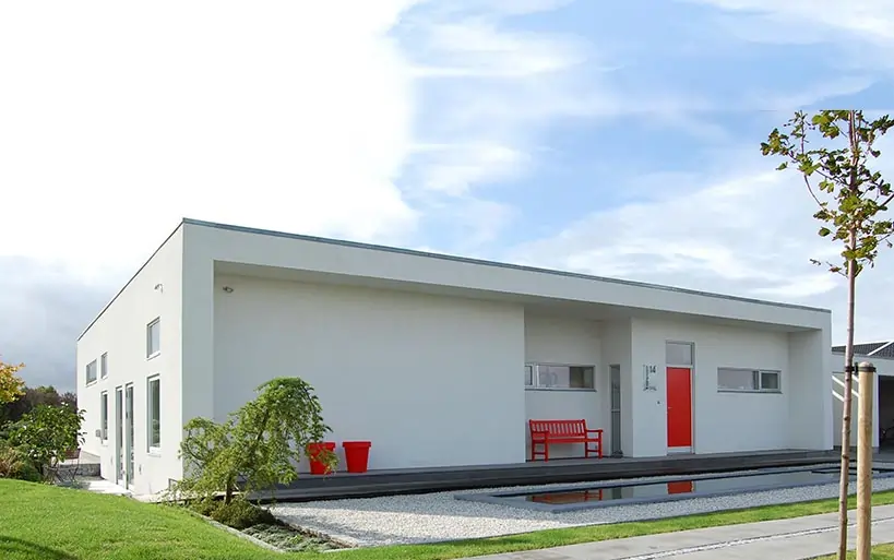 Weißer, moderner Flachdachbungalow mit minimalistischer Fassadengestaltung, roter Eingangstür, passender roter Bank und Pflanzgefäß im Vorgarten.