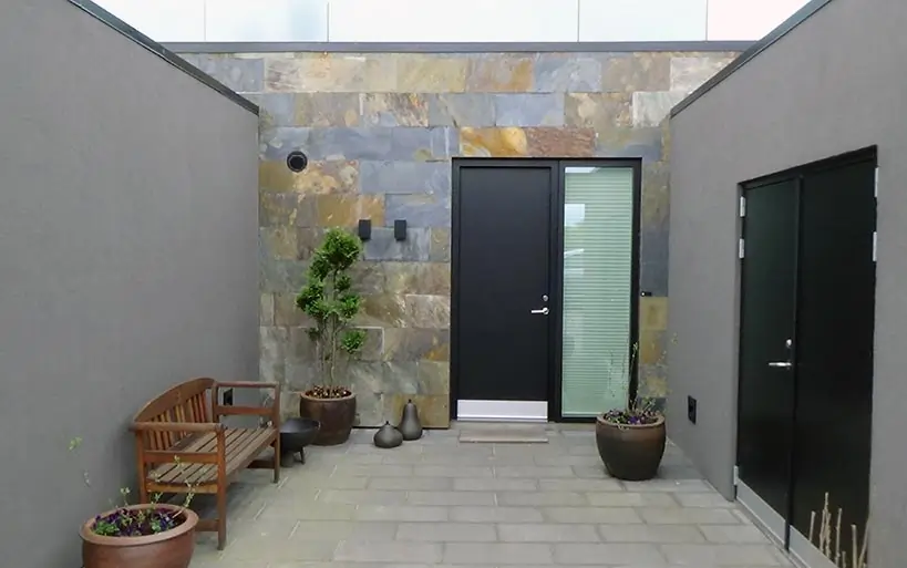 Moderne Hauseingangssituation mit Natursteinfassade, schwarzer Tür, seitlichem Fenster und dekorativen Elementen wie Bank und Pflanzkübeln.