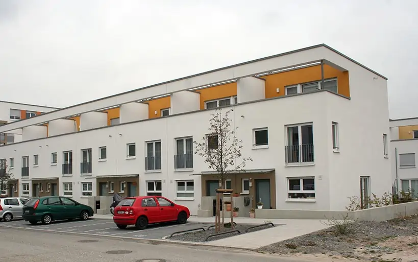 Moderne Reihenhausanlage mit weißen Fassaden, gelben Dachterrassen-Elementen und vorgelagerten Stellplätzen, aufgenommen an einem bewölkten Tag.
