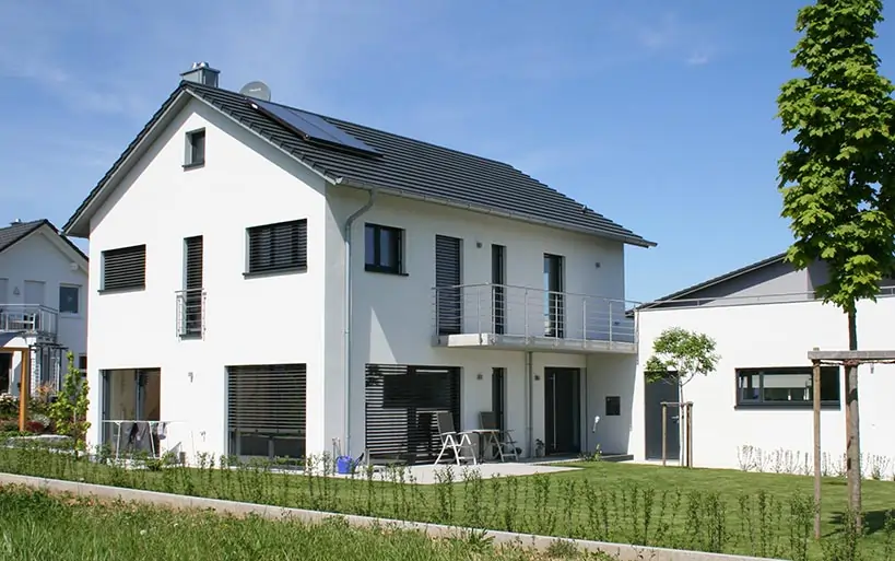 Modernes Einfamilienhaus mit weißer Fassade, grauem Satteldach und Balkon im Obergeschoss, daneben ein flacher Anbau und gepflegter Garten im Vordergrund.