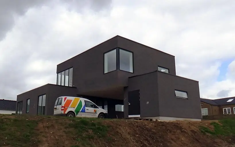 Modernes Flachdachhaus in Hanglage mit dunkler Fassade, großen Eckfenstern und einem geparkten Handwerkerfahrzeug davor.