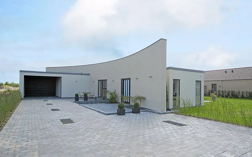 Modernes Einfamilienhaus mit geschwungenem Flachdach, heller Fassade, großer Einfahrt mit Pflastersteinen und gepflegtem Vorgarten.