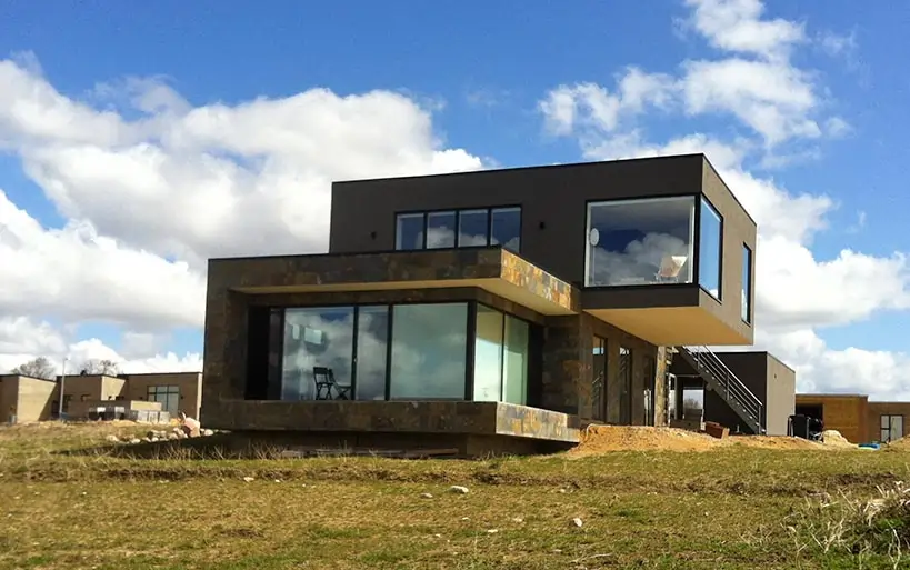Zweigeschossiges Flachdachhaus mit großen, bodentiefen Fenstern und moderner kubischer Architektur, erbaut auf einem leicht erhöhten Grundstück unter blauem Himmel.