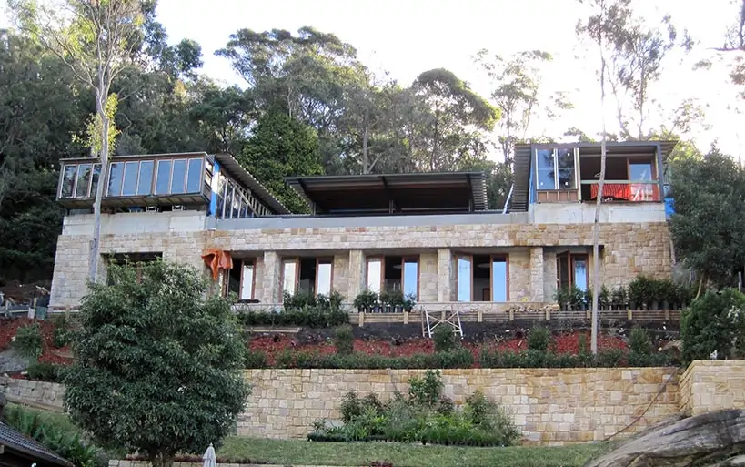 Modernes Terrassenhaus mit Natursteinfassade, großen Fenstern und Flachdach, eingebettet in eine bewaldete Hanglage mit aufwendig gestalteter Gartenanlage.