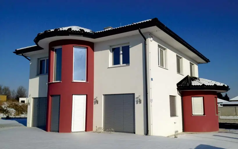 Modernes Zweifamilienhaus im Winter mit weißer Fassade, auffälligen roten Runderkern, flachem Walmdach und geschlossenen Rollläden.